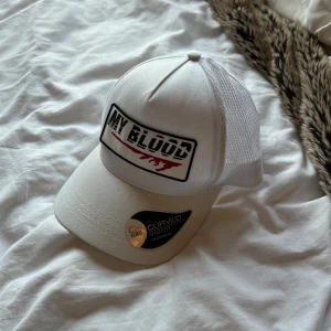Vit keps från Atlantis med tryck - Säljer en vit keps från Atlantis med curved visor och mesh på sidorna. Framtill finns ett broderat patch med texten 'MY BLOOD' och röd detalj. Kepsen har justerbar passform och är perfekt för dig som gillar streetwear.