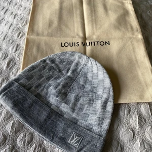 LV Mössa grå/silver - Exklusiv grå mössa från LV med klassiskt rutigt mönster och LV-logga framtill. Mössan är stickad i mjukt material och har en uppvikt kant. Perfekt accessoar för att lyfta din vinterstil.