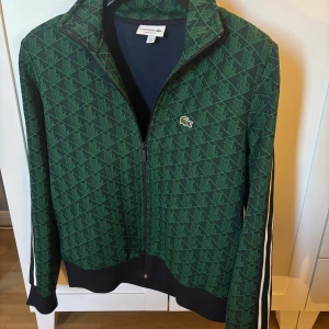 Grön mönstrad trackjacka Lacoste - Snygg grön trackjacka från Lacoste med geometriskt mönster och klassisk krokodillogga på bröstet. Jackan är i helt perfekt skick och använd fåtal gånger. För dig med skön o chill stil passar denna jacka perfekt.