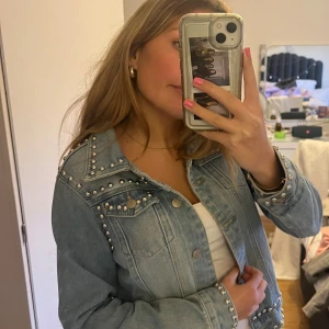 Ljusblå jeansjacka med nitar - Säljer en ljusblå jeansjacka med coola silverfärgade nitar på axlar, krage, ärmslut och nederkant. Jackan har klassiska bröstfickor med knappar och en snygg, rak passform. Perfekt för dig som vill ha en edgy och trendig look. Obs! Hålet på armen är inte en defekt utan de ska vara så! 