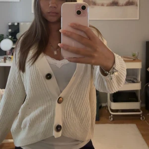 Vit stickad kofta med stora knappar - Säljer en vit stickad kofta med bred ribbad kant och tre stora kontrasterande knappar framtill. Koftan har en djup v-ringning och långa ärmar med ribbade muddar. Perfekt att slänga över en topp för en chill look.