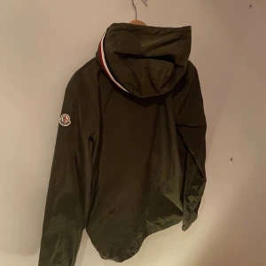 Grön vindjacka från Moncler - Snygg grön vindjacka från Moncler med huva och coola rödvita detaljer på kragen. Jackan har dragkedja framtill, snörning vid huvan och två fickor med knappstängning. Perfekt för dig som gillar stilren design och streetwear-vibe.