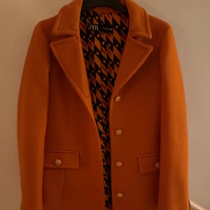 Orange kappa från Zara med guldknappar - Snygg orange kappa från Zara med klassisk krage och dekorativa guldknappar framtill. Fodret har ett coolt grafiskt mönster i svart och orange. Perfekt för dig som vill sticka ut med färg och stil i höst och vinter.