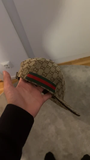 Gucci keps med GG-mönster och läderdetalj - Säljer en beige keps från Gucci med klassiskt GG-monogram i brunt över hela kepsen. Justerbar rem i brunt läder baktill och ikoniskt grönt/rött band på sidan. Kepsen är tillverkad i canvas och har snygga detaljer som ger en lyxig känsla.