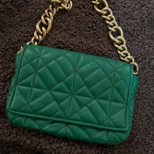 Grön quiltad axelväska från Zara - Snygg grön axelväska från Zara med quiltad design och grov guldfärgad kedja som axelrem. Väskan har magnetlås och ett rymligt fack med svart innerfoder. Perfekt storlek för det viktigaste och ger en cool touch till din outfit.