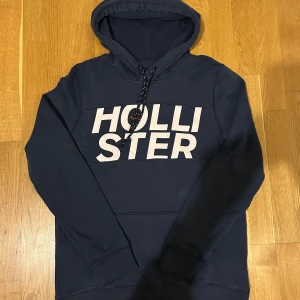 Mörkblå hoodie från Hollister - Snygg mörkblå hoodie från Hollister med stor vit logga på bröstet. Tröjan har huva med snörning och långa ärmar. Tillverkad i mjuk bomullsmix som är skön att ha på sig. Perfekt för chill dagar och när du vill ha en casual look.