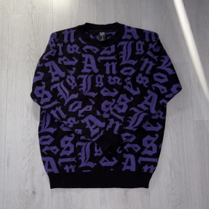 Svart och lila stickad tröja Black Squad - Cool stickad tröja från Black Squad i svart med lila grafiskt bokstavsmönster. Relaxed fit med ribbade muddar vid ärmslut och nederkant. Perfekt för dig som gillar streetstyle och vill sticka ut med ett unikt print.
