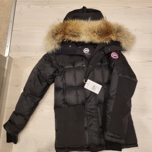 Svart dunjacka Canada Goose med pälskrage - Säljer en svart dunjacka från Canada Goose med stor huva och lyxig pälskant. Jackan har klassisk patch på ärmen, stora fickor och quiltad design. Perfekt för kalla dagar och riktigt snygg streetstil.