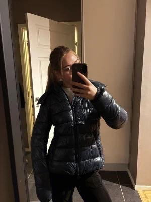Svart pufferjacka med dragkedja - Snygg mörkblå pufferjacka med glansig finish. Jackan har quiltad design och stängs med dragkedja framtill💥