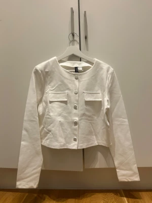 Vit croppad overshirt - Stilren vit croppad overshirt med rund halsringning, långa ärmar och dekorativa knappar framtill. Två fejkade bröstfickor ger en clean look. Perfekt för lager-på-lager och enkel att styla till olika outfits.