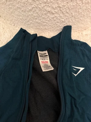 Mörk grön träningsjacka från Gymshark XS - Säljer en mörk grön långärmad träningsjacka från Gymshark i storlek XS. Jackan har dragkedja framtill, diskreta ventilationshål längs ärmarna och en sportig look med Gymshark-logga på bröstet. 