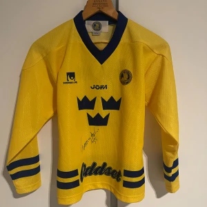 Svenska landslaget hockeytröja Salo #35 - Gul hockeytröja från svenska landslaget med blå detaljer, nummer 35 och namnet Salo på ryggen. Tröjan har långa ärmar, blå ränder på ärmar och nederkant samt Tre Kronor-tryck framtill. Materialet är mesh i polyester. Signerad av Tommy Salo.