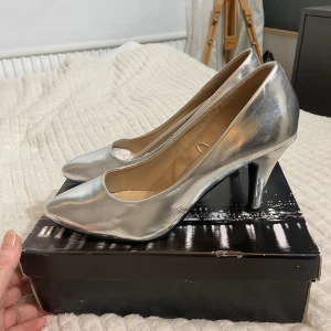 Silvriga pumps från SthlmDG, storlek 40 - Snygga pumps i metallic silver från SthlmDG. Skorna har spetsig tå och hög klack, vilket ger en riktigt trendig look. Tillverkade i syntetmaterial med en slät finish. Perfekta för dig som vill sticka ut med ett par glansiga klackar. Använd 1 gång har en liten skavanker som syns på bilden.