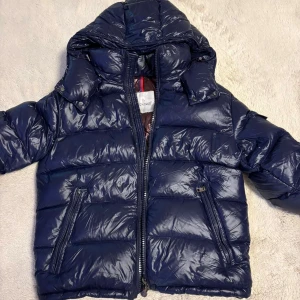 Mörkblå Moncler pufferjacka med huva - Säljer en mörkblå pufferjacka från Moncler med glansig finish och avtagbar huva. Jackan har två snedställda fickor med dragkedja, quiltad design och ett klassiskt Moncler-märke på ärmen. Insidan är brun och har en cool serietidningsdetalj. Nfc funkar🫡