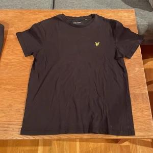 Svart t-shirt från Lyle & Scott M - Svart t-shirt från Lyle & Scott i bomull med klassisk passform. T-shirten har korta ärmar och pryds av den ikoniska gula örnloggan broderad på bröstet. Enkel och stilren design som funkar till det mesta.