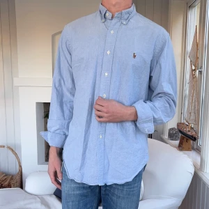 Blå skjorta från Ralph Lauren - Klassisk ljusblå skjorta från Ralph Lauren med button-down krage och broderad logga på bröstet. Skjortan har lång ärm, normal passform och är tillverkad i mjuk bomull. Perfekt för en clean och stilren look.