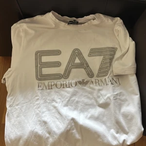 Vit EA7 Emporio Armani t-shirt - Vit t-shirt från EA7 Emporio Armani med stor logga och tryck framtill. Klassisk rund hals och korta ärmar. Tillverkad i mjuk bomull för en skön känsla. Perfekt för dig som gillar stilrena och exklusiva plagg med tydlig branding.