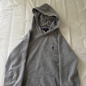 Grå hoodie från Polo Ralph Lauren - Säljer en klassisk grå hoodie från Polo Ralph Lauren med mörkblå broderad logga på bröstet. Tröjan har huva med snörning och en stor magficka. Mjuk insida och långärmad modell, perfekt för chill dagar.