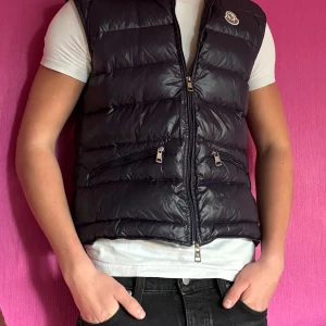 Marinblå dunväst från Moncler - Snygg marinblå dunväst från Moncler. klassisk Moncler-logga på bröstet och tvåvägsdragkedja. Perfekt för lager-på-lager. Går att dra upp dragkedjan smidigt ändå om den är lite trasig som visas på bild 4. Storlek 1 vilket motsvarar storlek S.