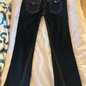 True Religion Joey Low Rise Flare Jeans - Snygga svarta jeans från True Religion, modell Joey Low Rise Flare. De har låg midja och utsvängda ben, med kontrasterande vita sömmar och detaljerade fickor bak. Klassisk knappstängning och coola broderade detaljer som ger extra edge.