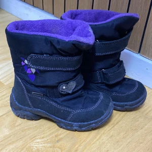 Svarta vinterstövlar med lila foder - Svarta vinterstövlar med mjukt lila fleecefoder och kardborreband. Dekorativa hjärtan på sidan och glittrig detalj på remmen. Grov sula för bra grepp och förstärkta partier i mocka. Perfekta för kalla dagar.