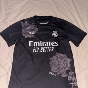Real Madrid Y-3 svart fotbollströja - Säljer en svart Real Madrid Y-3 fotbollströja med vita tryck, stora rosor som detaljer och Bellingham 5 på ryggen. Tröjan har korta ärmar och är tillverkad i ett lätt, ventilerande material. Perfekt för dig som vill sticka ut på planen eller läktaren.