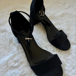 Svarta klackar från XIT - Stilrena svarta klackar från XIT med bred rem över tårna och justerbart ankelband med silverfärgat spänne. Skorna har blockklack och öppen tå, tillverkade i mockaliknande material som ger en mjuk känsla. Lätta att gå i och bekväma. Använda fåtal gånger.