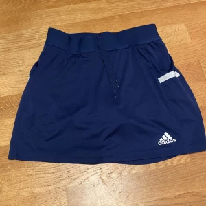 Adidas - Mörkblå sportkjol - Badminton/Tennis  - Sportig mörkblå kortkjol från Adidas med inbyggda shorts och elastisk midja med snörning. Kjolen har vita detaljer på sidorna och Aeroready-tyg som andas. Perfekt för tennis eller träning, stilren och bekväm design.