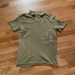 Olivgrön pikétröja från H&M - Olivgrön pikétröja från H&M med klassisk krage och knappar framtill. Tröjan har korta ärmar och en liten broderad logga på bröstet. Perfekt för en avslappnad och stilren look.