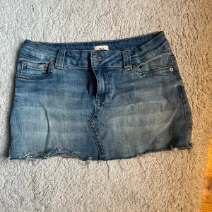 Blå jeanskjol från Nelly - Snygg blå jeanskjol från Nelly med klassisk femficksdesign och rå, fransig nederkant. Kjolen har bälteshällor och dragkedja framtill. Perfekt för en avslappnad och trendig look.