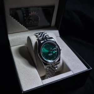 Seiko Mod Green Sunburst Klocka - Säljer en stilren Seiko klocka med automatisk urverk, green sunburst urtavla med arabiska siffror och datumvisning samt luminous handvisare. 39 mm klockan med ett klassiskt rundat silverfärgat boett och länkarmband i 904L stäl. Glas på båda sidorna gjort av safir vilket innebär att den är reptålig. Perfekt accessoar för dig som gillar snygga detaljer och modern design.