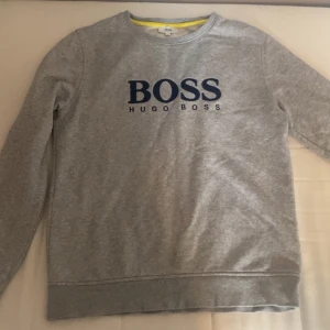 Grå sweatshirt från Hugo Boss - Grå sweatshirt från Hugo Boss med mörkblå BOSS-logga framtill. Klassisk rund halsringning och långa ärmar. Tillverkad i mjuk bomullsmix, perfekt för en chill och stilren look.