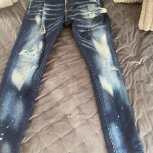 Dsquared2 blå distressed jeans - Säljer ett par blå jeans från Dsquared2 med coola slitningar, blekta partier och färgstänk. Byxorna har rak passform, klassiska fem fickor och unika detaljer som broderad text på bakfickan och stor patch med 'Cool Bella' bak. Perfekta för dig som gillar streetstyle.