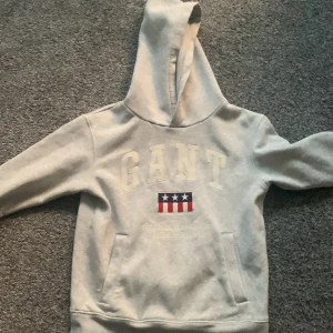 Grå hoodie från GANT med tryck - Ljusgrå hoodie från GANT med broderad logga och amerikanskinspirerat tryck framtill. Tröjan har huva, känguruficka och långa ärmar. Mjuk insida och klassisk passform, perfekt för chill dagar.