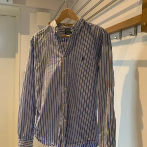 Randig skjorta från Polo Ralph Lauren - Snygg randig skjorta från Polo Ralph Lauren i blått och vitt med broderad logga på bröstet. Skjortan har en slim fit passform och är långärmad med knappar framtill. Perfekt för en stilren look. ( pris går att diskutera)