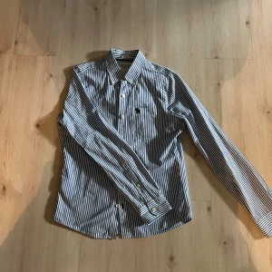 Randig skjorta från Abercrombie & Fitch - Snygg skjorta från Abercrombie & Fitch i vitt och blått med klassiska vertikala ränder. Skjortan har lång ärm, knappar framtill och en liten broderad älg på bröstfickan. Tillverkad i bomull och har en normal passform.