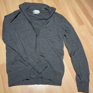 Grå merinoullskofta från These Glory Days - Mörkgrå kofta i mjuk merinoull från These Glory Days. Klassisk modell med dragkedja framtill, ribbade muddar och lång ärm. Perfekt för lager-på-lager och snygg till både jeans och chinos.