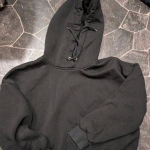 Svart kort hoodie med snörning st xs - En klassisk svart hoodie med snörning i huvan och ribbade muddar vid ärmsluten. Perfekt för chill dagar eller när du vill ha något bekvämt och enkelt. Tillverkad i mjuk bomullsmix och har en avslappnad passform. En kortare modell