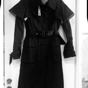 Svart trenchcoat från L'Ecole National XS - Snygg svart trenchcoat från L'Ecole National i storlek XS. Jackan har dubbelknäppning, bälte i midjan, stora fickor och klassisk krage. Fodret är champagnefärgat med små prickar. Perfekt för dig som vill ha en stilren och tidlös look.