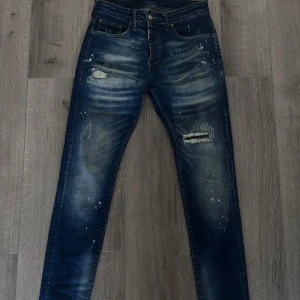 Dsquared2 byxor slim - Säljer ett par Dsquared2 jeans slim i mörkblå tvätt med slitningar, lagade hål och coola färgstänk. Jeansen har klassisk femficksdesign, orange sömmar och smal passform. Perfekta för dig som gillar en streetig och avslappnad look.