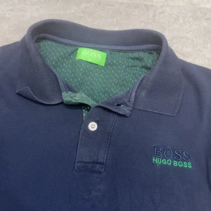 Mörkblå Hugo Boss pikétröja M - Snygg mörkblå pikétröja från Hugo öBoss med klassisk krage och två knappar framtill. Broderad logga i blått och grönt på bröstet. Insidan av kragen har ett grönt tyg med små mönsterdetaljer. Perfekt för en chill och stilren look.