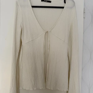 Vit ribbad blus från Bik Bok - Supersöt vit blus från Bik Bok med lång ärm och v-ringning. Toppen har ribbad struktur, knytning framtill och lätt vågiga kanter vid ärmslut och nederkant. Tillverkad i mjukt material med viskos och polyester för en skön känsla.