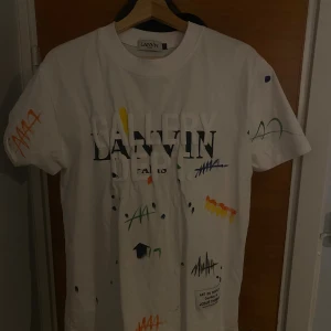 Vit Gallery Dept x Lanvin t-shirt - Vit t-shirt från Lanvin x Gallery Dept med färgstänk och graffiti-liknande detaljer i grönt, blått, gult, rött och orange. Stor tryckt text fram och bak, där baktexten är i guldglitter. Skön passform och cool streetkänsla. Perfekt statement-plagg. Säljer billigt då jag inte vet om den är äkta! Hör av er!