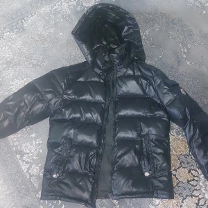 Svart pufferjacka från Moncler - Säljer en svart pufferjacka från Moncler med glansig finish och huva. Jackan har två fickor med dragkedja framtill och en Moncler-logga på ärmen. Perfekt för kalla dagar och riktigt snygg till streetwear-stilen.