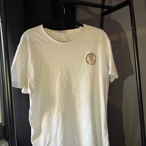 Vit Burberry t-shirt med logga - Snygg vit t-shirt från Burberry med klassisk rund hals och korta ärmar. På bröstet finns en liten Burberry-logga med rutigt mönster i typisk Burberry-stil. T-shirten är gjord i mjuk bomull och har en clean, stilren look. lite smutsig dock på lappen, men det syns inte