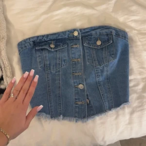 Blå jeans tubtopp med knappar - Säljer en snygg blå jeans tubtopp med knappar framtill och två dekorativa fickor. Toppen har fransad nederkant och smockad rygg för extra komfort. 