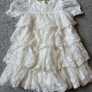 Vit spetsklänning från Newbie 86 - Superfin vit klänning från Newbie Limited Edition i storlek 86. Klänningen har flera lager volanger, broderade blommor och puffiga korta ärmar i tunn spets. Perfekt för speciella tillfällen och ger en riktigt söt look.