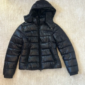 Svart pufferjacka med huva - Svart pufferjacka från lager 157 i storlek S. Jackan har en glansig finish, avtagbar huva och dragkedja framtill samt två snedställda fickor med dragkedja. Perfekt för kalla dagar och riktigt snygg till streetwear-stilen.