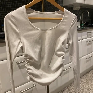 Vit ribbad långärmad topp från SHEIN - Säljer en vit långärmad topp från SHEIN med ribbad struktur och snygg rynkdetalj på sidorna. Toppen har rund halsringning och är figurnära i modellen. Perfekt att styla med jeans eller kjol för en clean look.