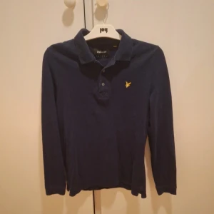 Mörkblå långärmad pikétröja Lyle & Scott - Snygg mörkblå långärmad pikétröja från Lyle & Scott med klassisk krage och knäppning framtill. Tröjan har det ikoniska gula logobroderiet på bröstet och är tillverkad i mjuk bomull. Aldrig använd, storlek S